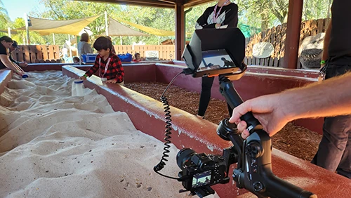 A child playing in the sand at a theme park being recorded by a video camera